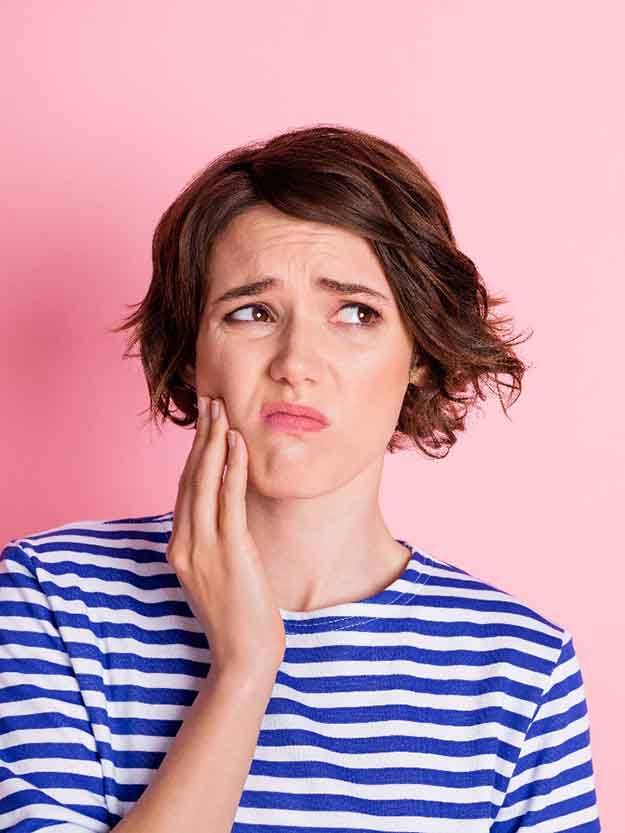 Woman with short brown hair and blue/white striped shirt holding her jaw in pain in front of pink background